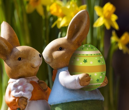 Zwei frühlingshafte Hasenfiguren halten ein buntes Osterei, umgeben von gelben Narzissen.