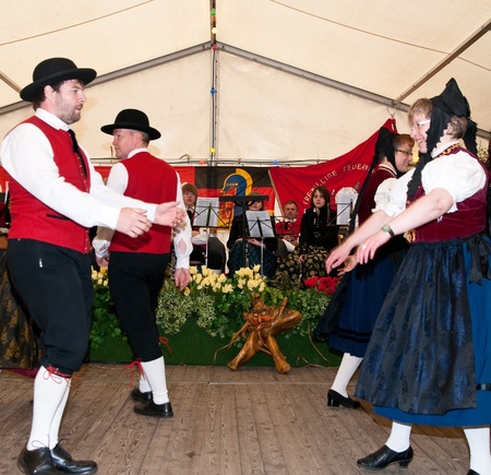 Tanzende Paare in traditioneller Kleidung auf einer Feier, im Hintergrund eine Band und dekorative Blumenarrangements.