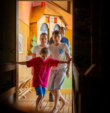 Vier Personen betreten einen Raum, in dem ein Spielhaus im Hintergrund zu sehen ist. Zwei Kinder und zwei Erwachsene sind ...