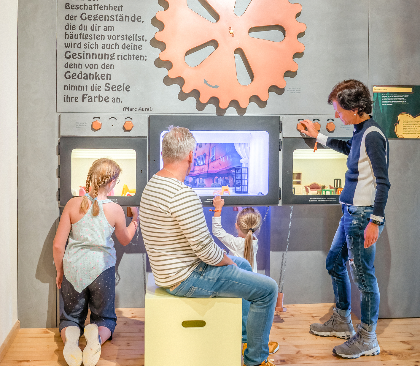 Familie interagiert mit einer interaktiven Ausstellung, Kinder schauen in Kästen, während Erwachsene anschaulich erklären.