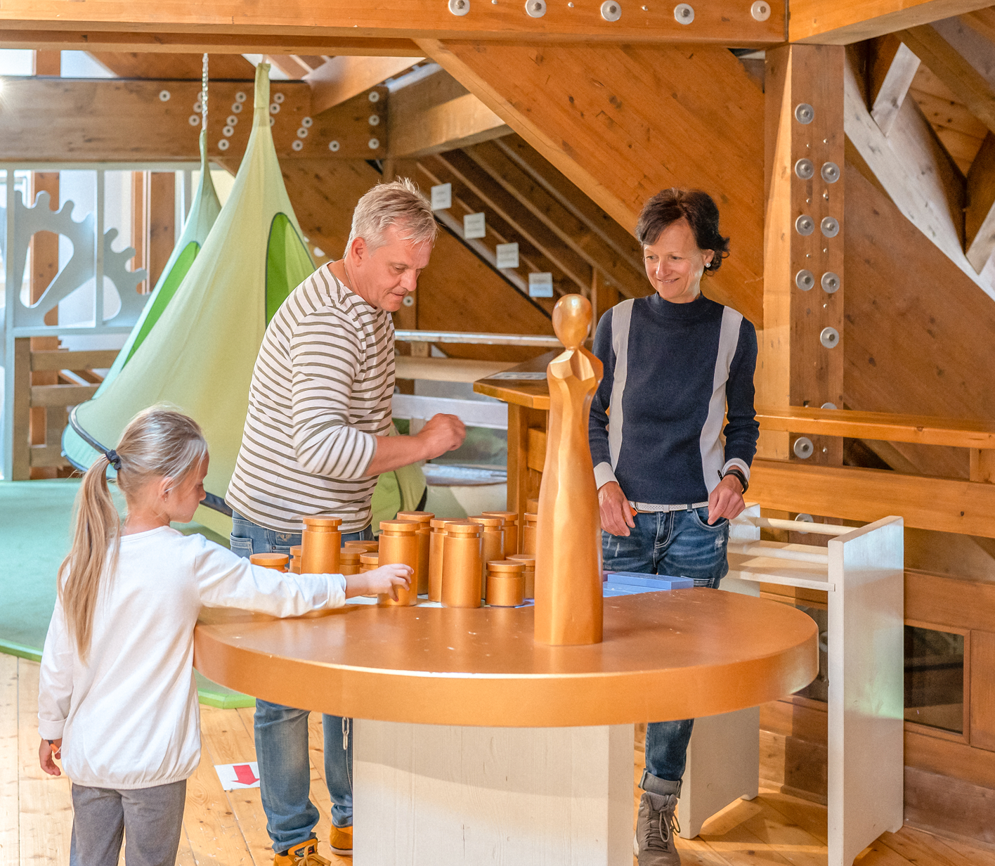 Eine Familie spielt mit Holzbehältern an einem Tisch in einem hellen Spielraum mit Holzelementen.