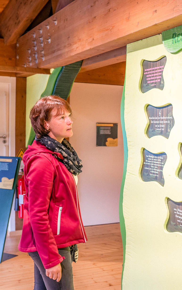 Eine Frau in roter Jacke betrachtet interaktive Ausstellungstafeln in einem Holzraum.