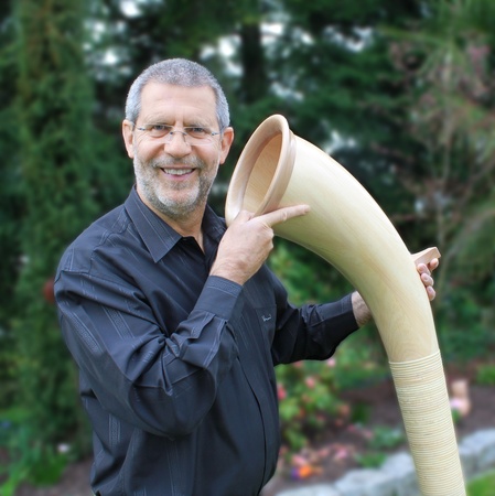 Mann in schwarzer Bluse hält ein großes Musikinstrument und lächelt, umgeben von grüner Gartenlandschaft.