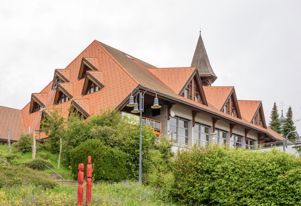 Haus mit rotem Satteldach, großen Fenstern und Türmen, umgeben von grünem Gelände und Skulpturen.