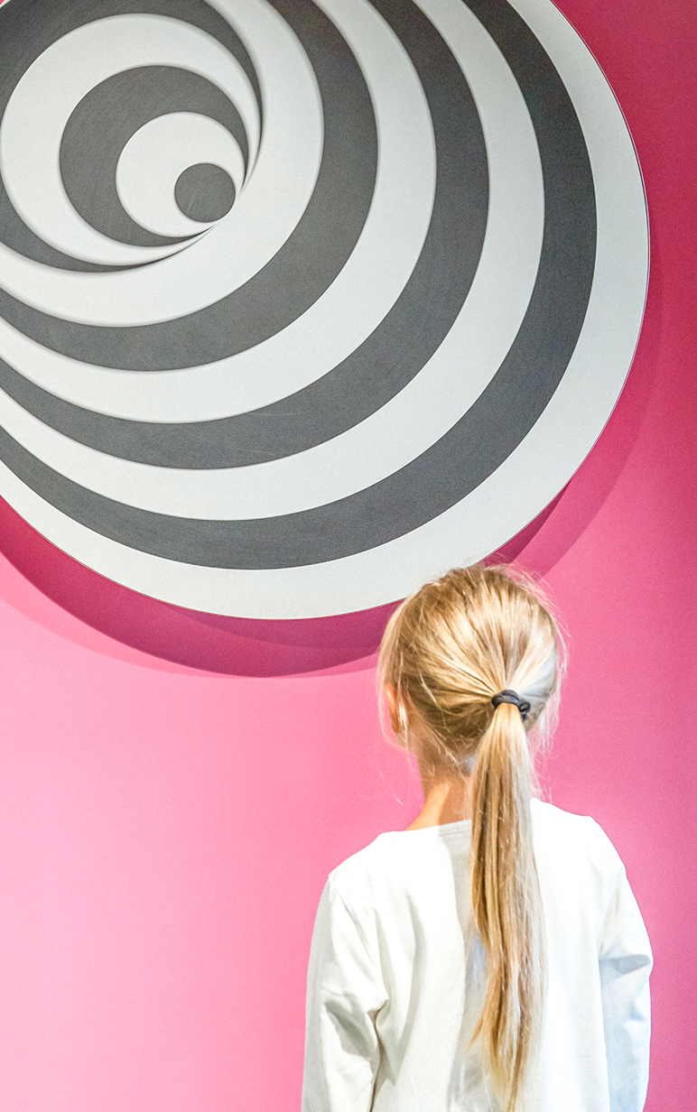 Ein Kind steht vor einer spiralförmigen Wandinstallation in Schwarz-Weiß, vor pinker Hintergrundfarbe.