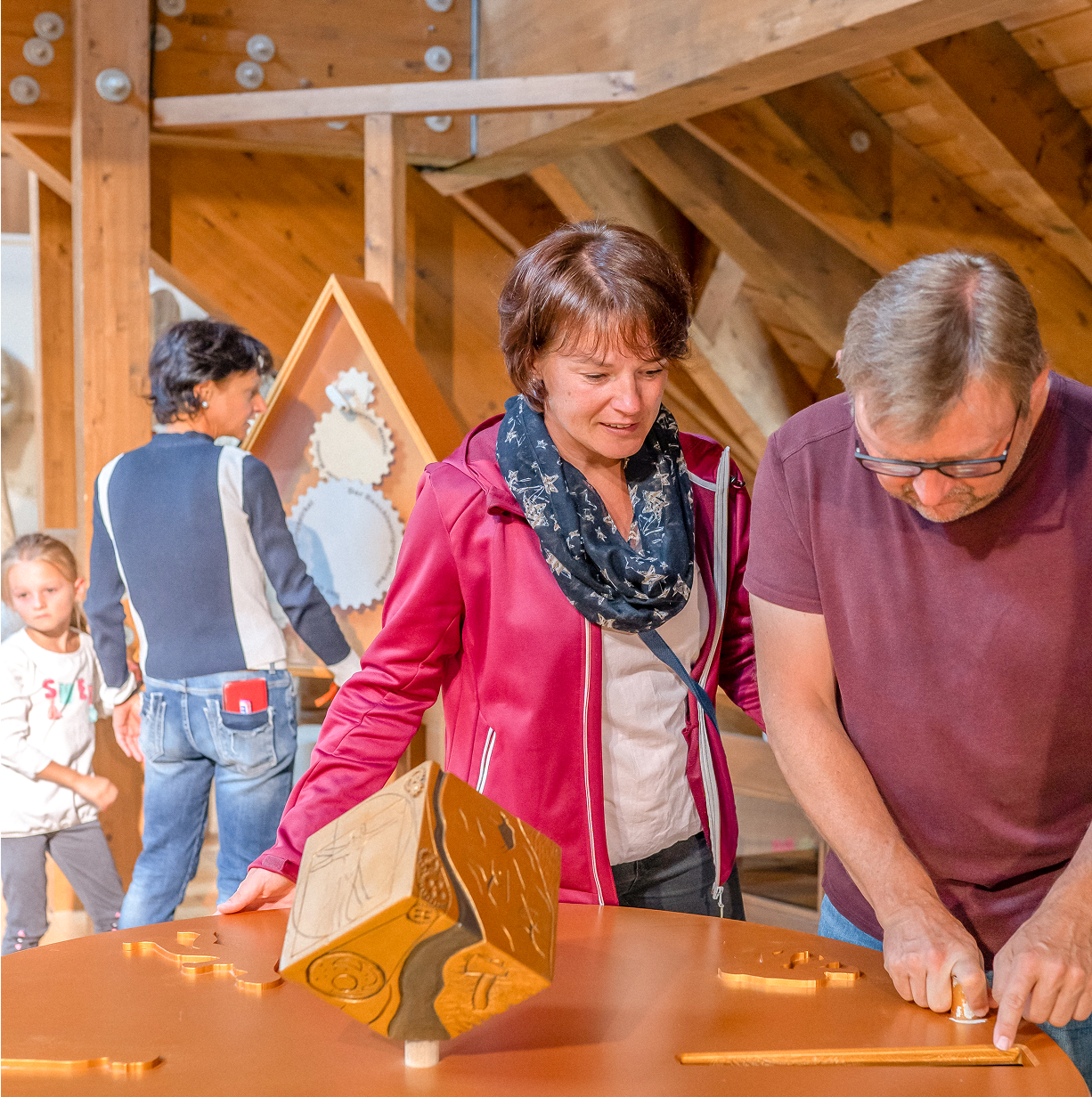 Eine Frau und ein Mann untersuchen einen Holzwürfel, während weitere Besucher im Hintergrund eine Ausstellung erkunden.