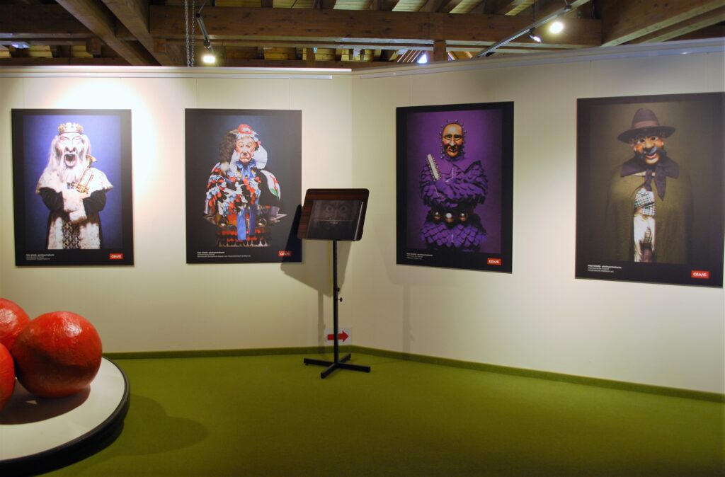 Eine Kunstinstallation mit vier Fotos von maskierten Figuren an einer Wand und einer Stütze im Vordergrund.