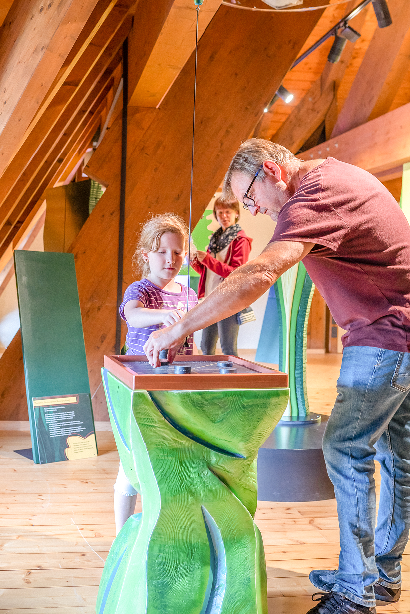 Ein Mann zeigt einem Kind eine interaktive Ausstellung, während eine Frau im Hintergrund zuschaut. Holzinterieur sichtbar.