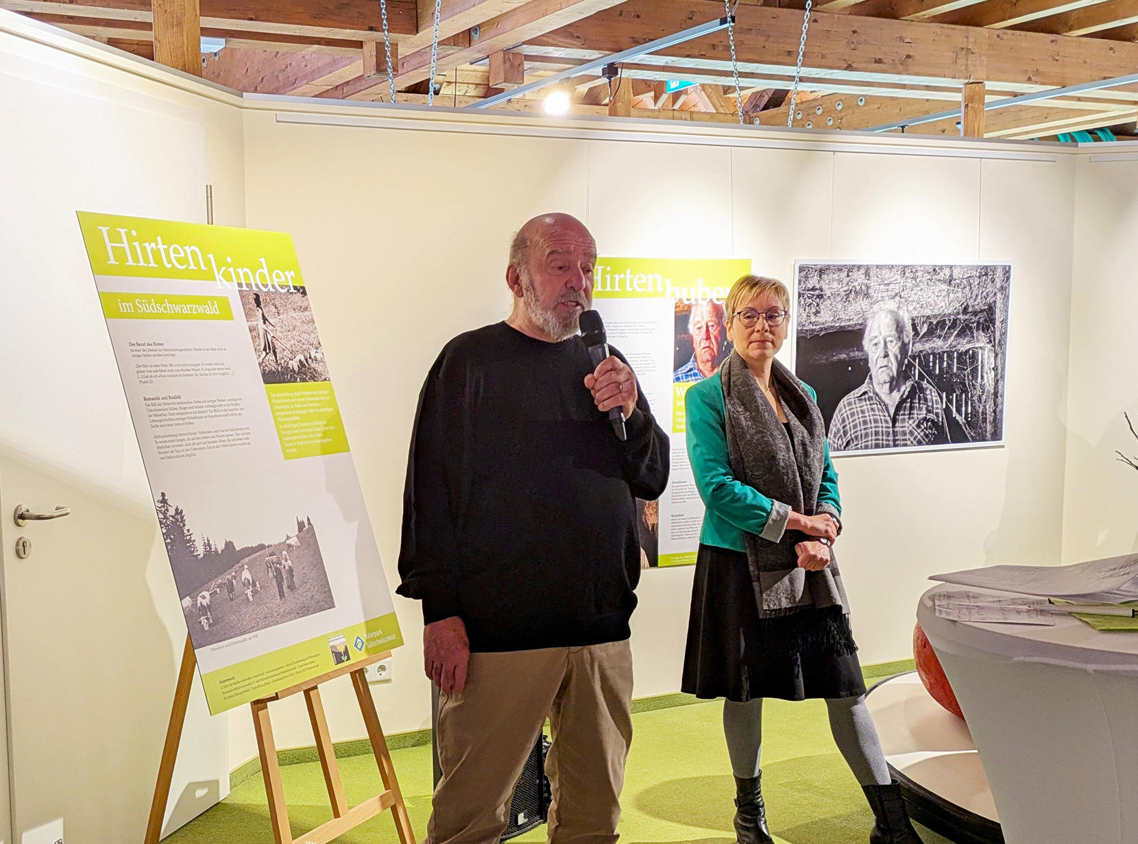 Zwei Personen sprechen bei einer Ausstellung über Hirtenkinder im Südschwarzwald, Umgebungsinfos im Hintergrund.