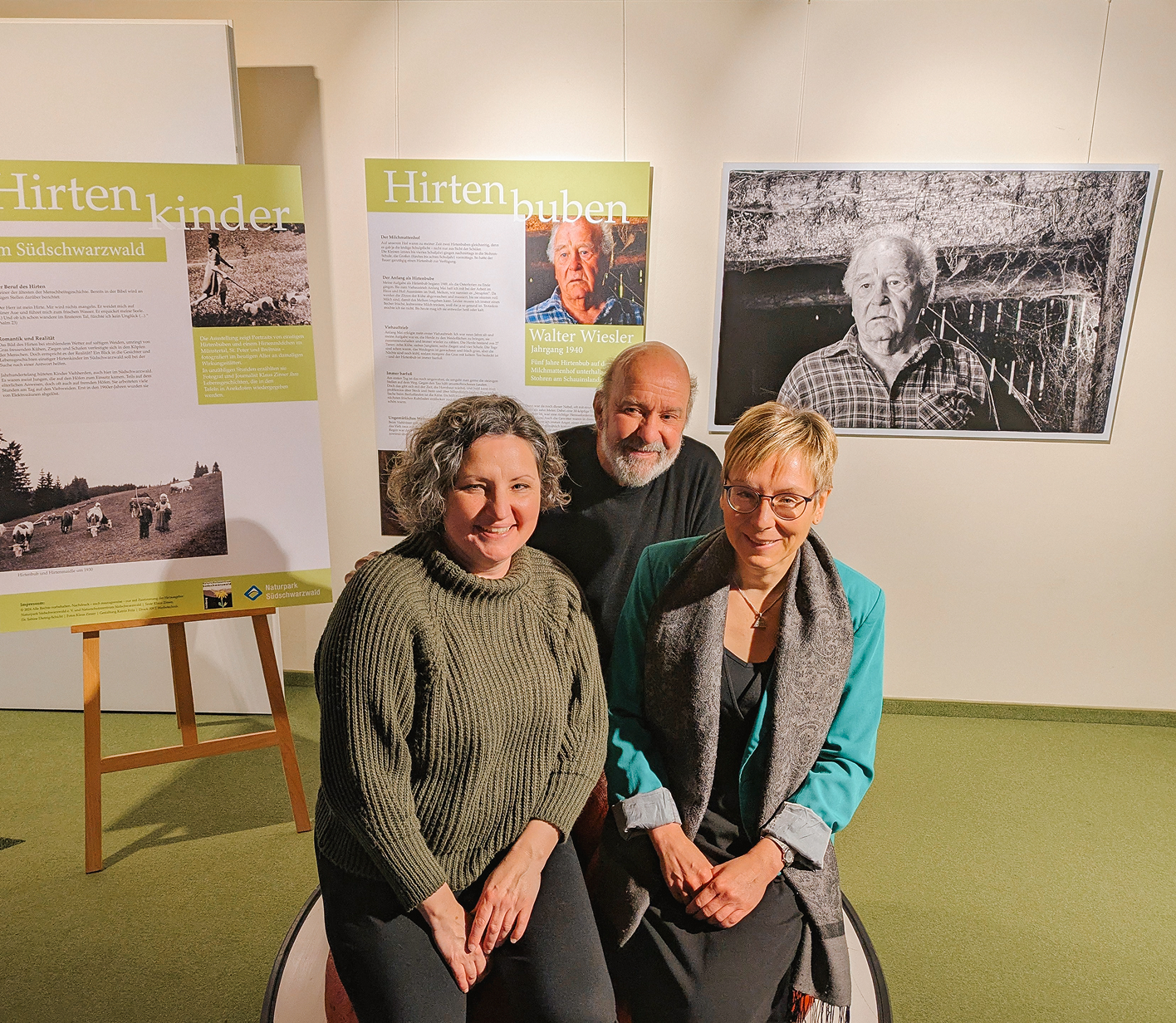 Drei Personen posieren lächelnd vor großformatigen Bildern und Informationsplakaten in einer Ausstellung.
