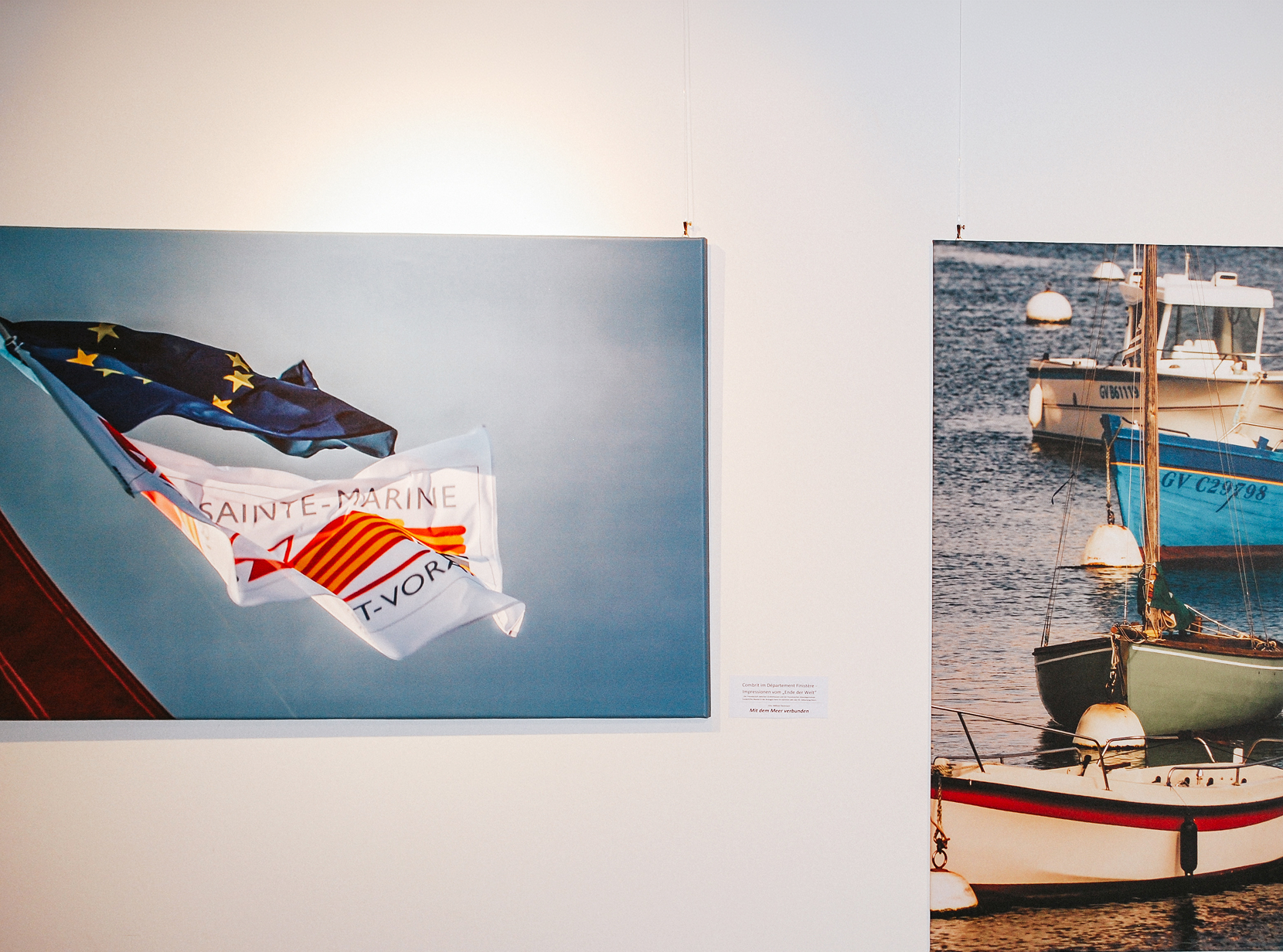Zwei Bilder in einer Ausstellung: Eine Flagge weht, daneben ein Hafen mit Booten im Wasser.