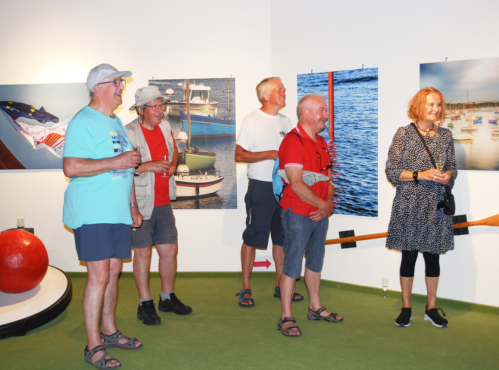 Fünf Personen stehen in einer Galerie und betrachten Bootsfotografien an den Wänden. Zwei halten Getränke.