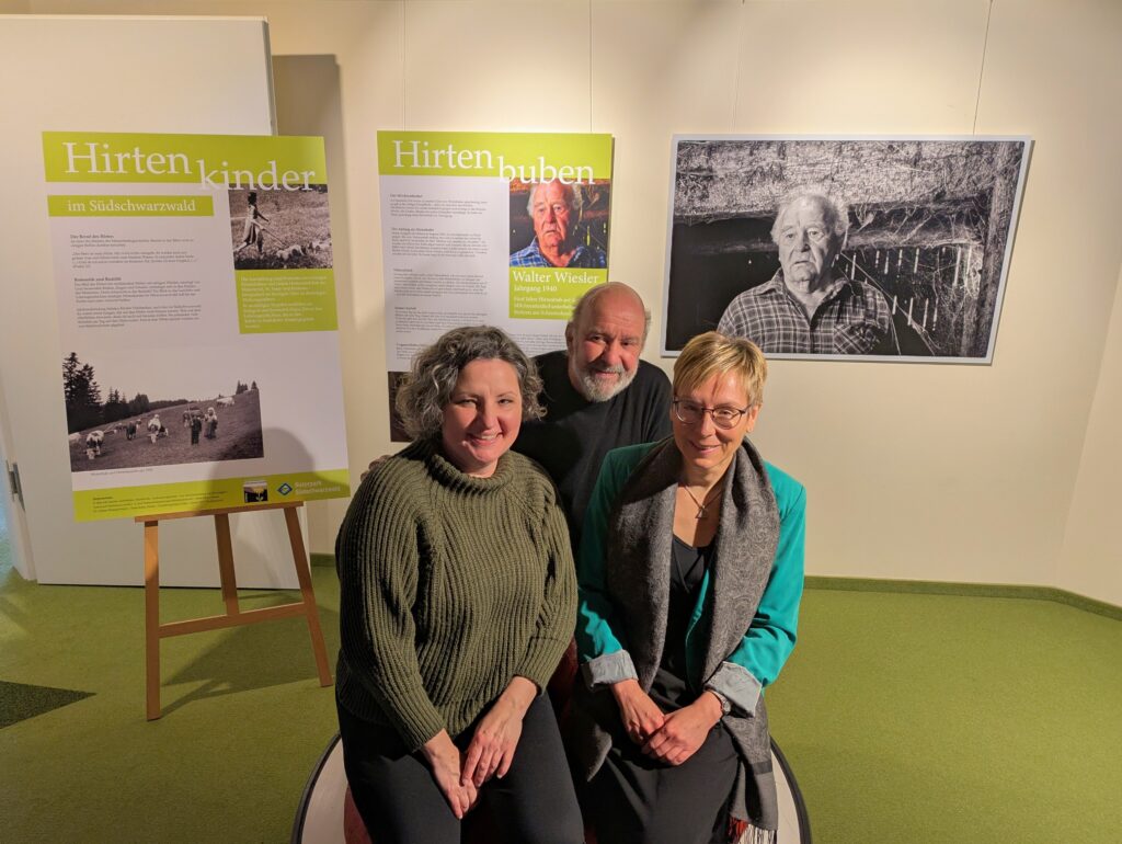 Drei Personen sitzen im Vordergrund einer Ausstellung, hinter ihnen Informationstafeln über Hirten im Südschwarzwald.