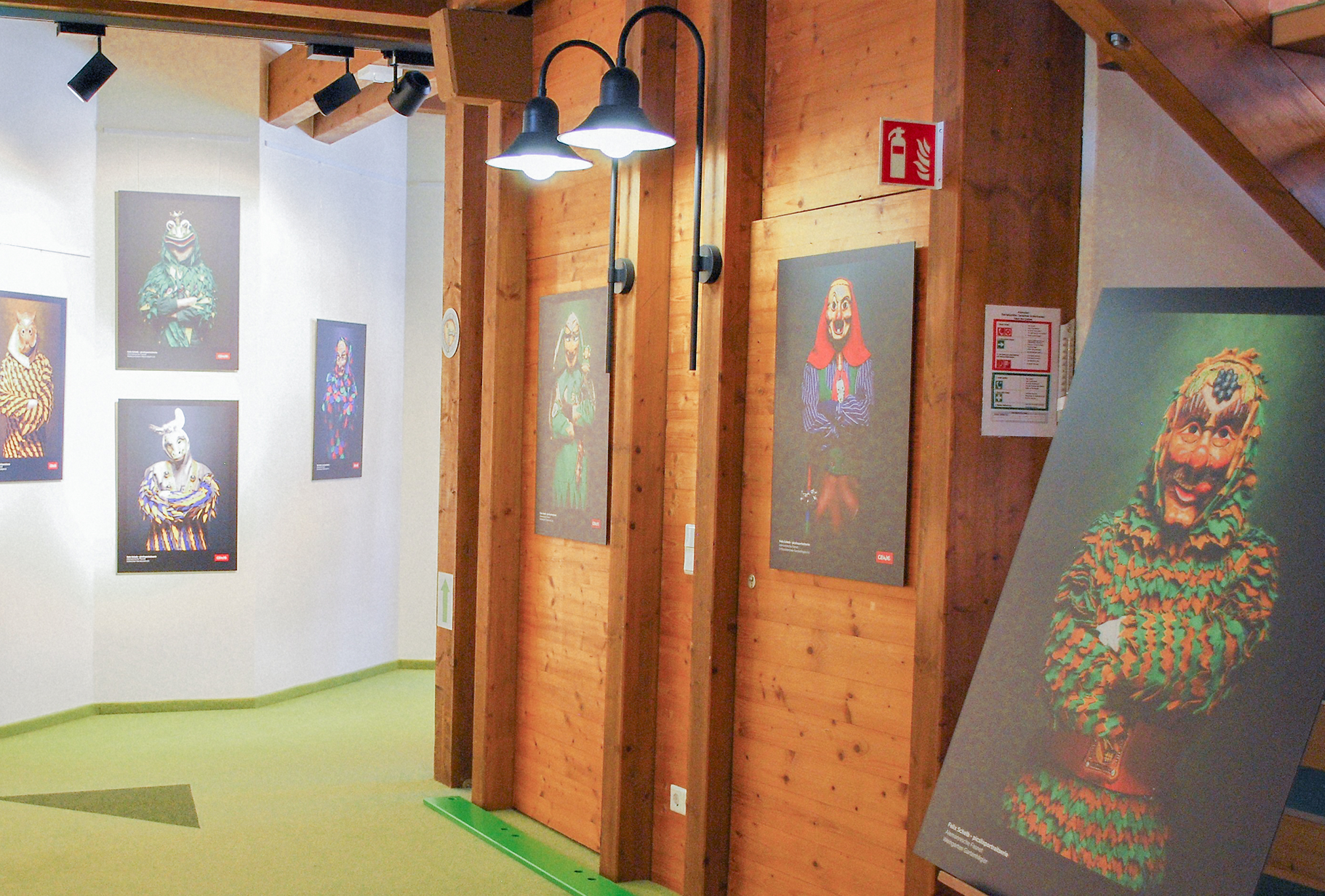 Ein Ausstellungsraum mit Bildern von maskierten Figuren an Holzwänden und beleuchtet von zwei Lampen.