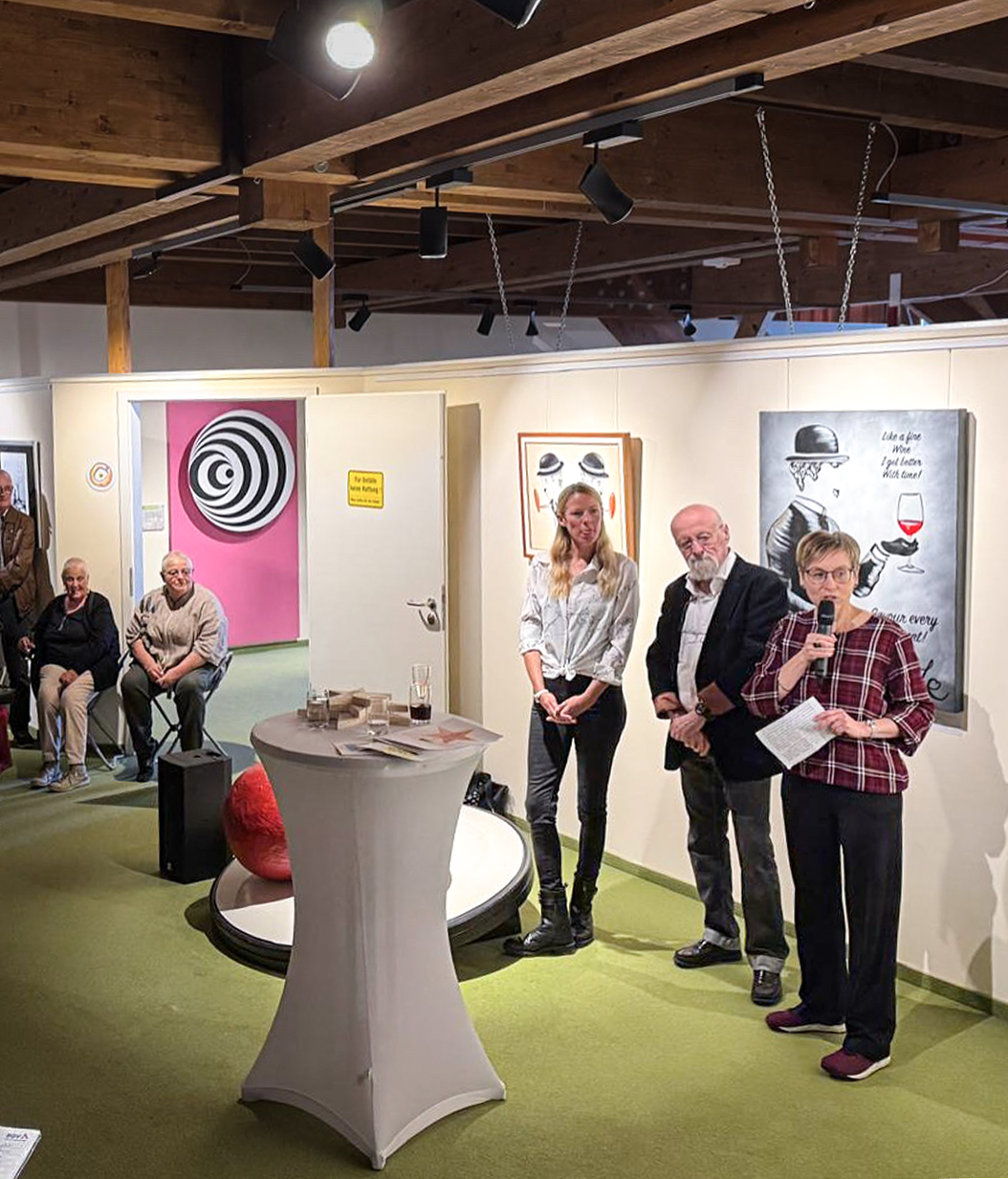 Eröffnung einer Kunstausstellung mit mehreren Personen, die sprechen, und Zuschauern im Hintergrund.